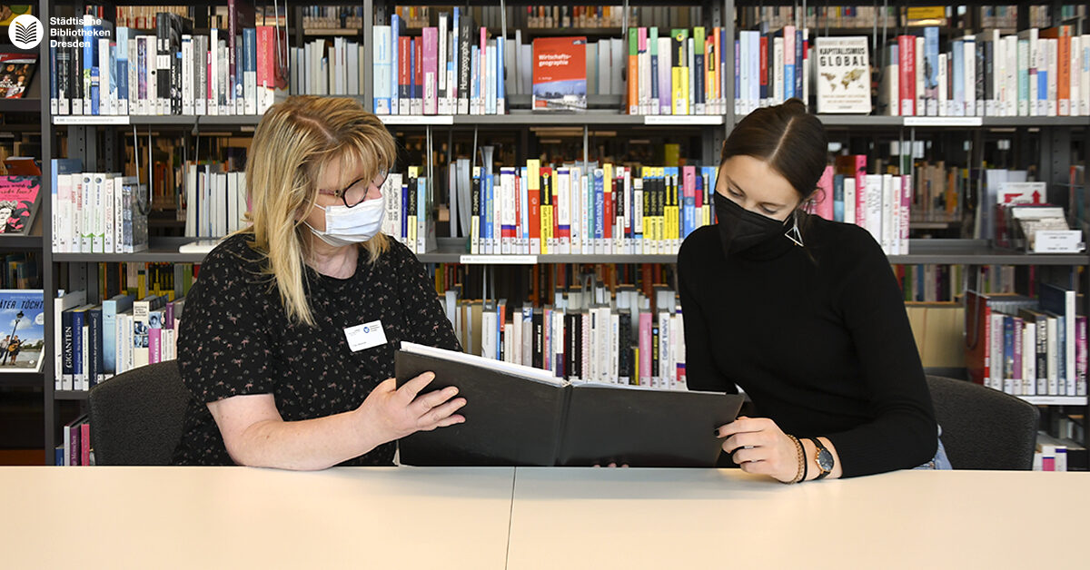 Zwei Frauen sitzen an einem Tisch und schauen in eine Mappe.