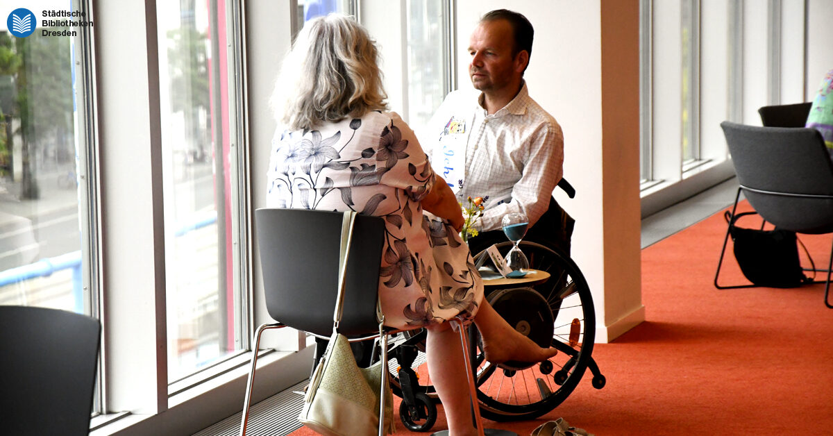 Zwei Personen unterhalten sich im Foyer der Zentralbibliothek. Zwischen ihnen befindet sich ein Tisch mit einer Sanduhr.