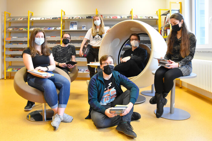 Das Foto zeigt drei jungen Frauen und drei jungen Männern in der Bibliothek. Sie tragen Mund-Nasen-Schutz und halten verschiedene Bibliotheksmedien in der Hand.