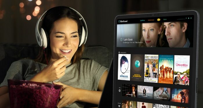 Eine Frau sitzt mit Kopfhörern auf dem Sofa und schaut auf ihr Tablet, welches ihr verschiedene Filme vorschlägt.