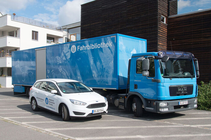 Foto: Zugmaschine mit Sattelauflieger der Fahrbiblothek, davor ein PKW des Bücherhausdienstes