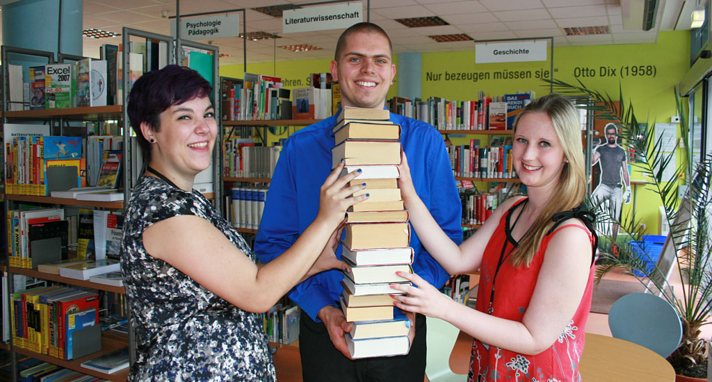 Drei Auszubildende (links und rechts eine Frau, in der Mitte ein Mann) halten gemeinsam einen Stapel Bücher Drei Auszubildende (links und rechts eine Frau, in der Mitte ein Mann) halten gemeinsam einen Stapel Bücher