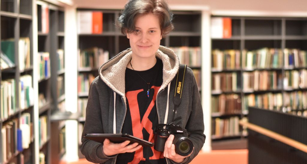 Eine junge Frau mit Kamera und Tablet steht vor einem Bücherregal. Eine junge Frau mit Kamera und Tablet steht vor einem Bücherregal.