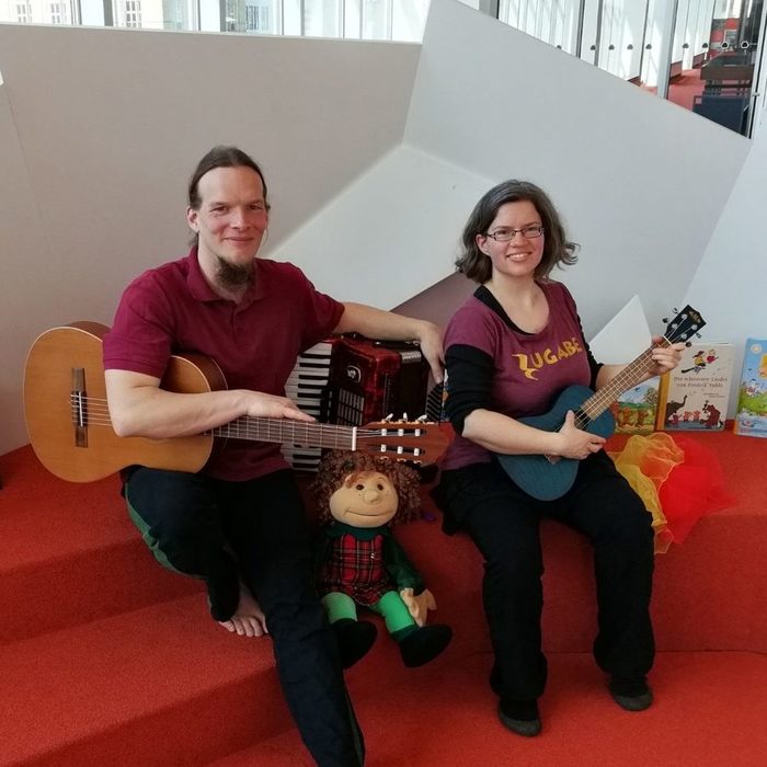 Zwei Musiker*innen sitzen in der Kinderbibliothek und halten Musikinstrumente in der Hand.