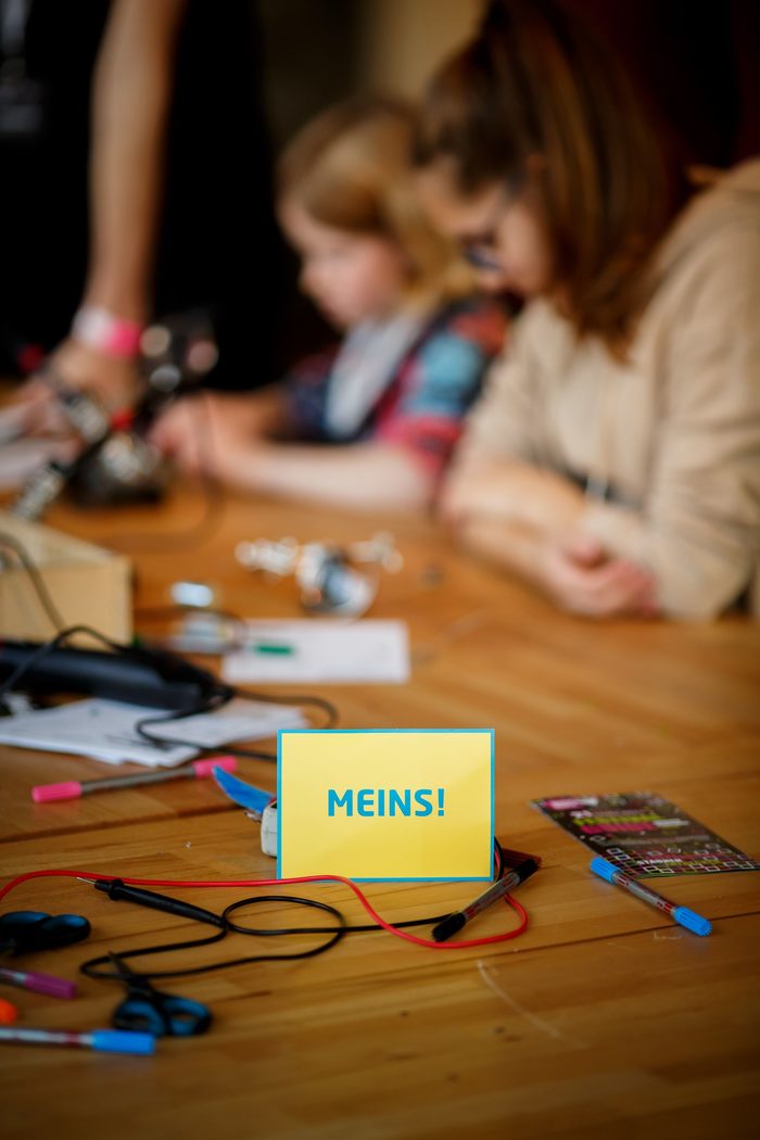 Eine Box mit der Aufschrift "MaKey" ist im Vordergrund zu sehen. Im Hintergrund liegen Löffel, welche mit elektrischen Kabeln mit der Box verbunden sind.