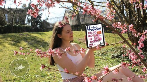 Person mit dem Buch "Nur noch ein einziges Mal" sitzt neben einem blühenden Kirschbaum