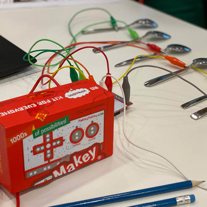 Eine Box mit der Aufschrift "MaKey" ist im Vordergrund zu sehen. Im Hintergrund liegen Löffel, welche mit elektrischen Kabeln mit der Box verbunden sind.