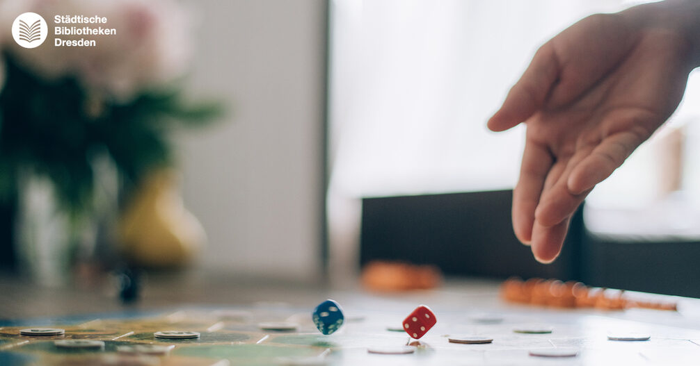 Close-Up von einer Hand, die Würfel auf ein Brettspiel wirft.